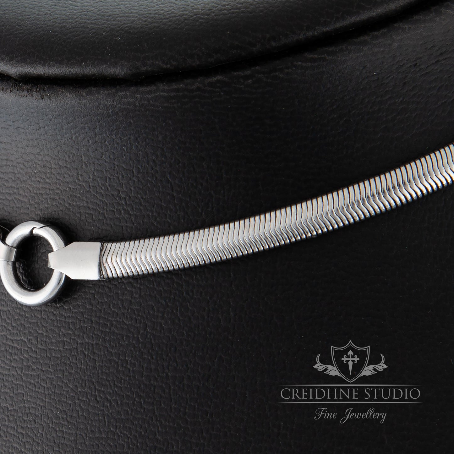 Close-up of a silver snake chain on a black leather surface with 'Creidhne Studio' branding.